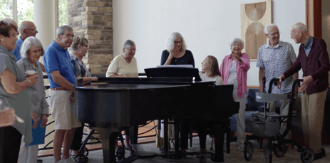 A group of elderly adults around a grand piano in a room with light walls and a stone pillar.