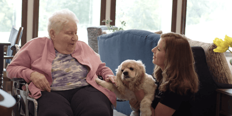 An elderly woman in a wheelchair interacts with a young woman holding a small dog in a bright room.