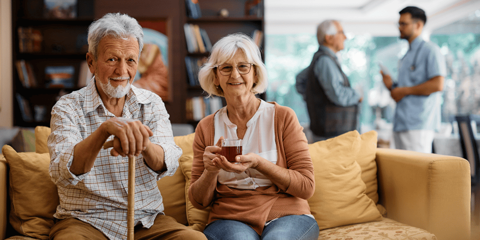 A smiling senior couple sitting on a couch. The man is holding a cane, and the woman has a cup of tea.